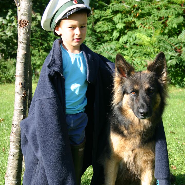 Kind in einem großen Mantel und mit Uniformmütze steht neben einem Schäferhund